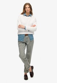 Jonge vrouw met lang haar, gekleed in een wit sweatshirt over een blauwe spijkerblouse, gestreepte broek en donkere instapschoenen, staand met de armen gekruist.