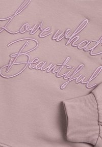 Světle růžový svetr s výšivkou "Miluj to, co je krásné." Má žebrované manžety a měkkou strukturu.