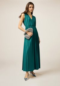 Türkise plissierte Maxikleid mit V-Ausschnitt und Taillenbund, kombiniert mit einer silbernen Metallic-Clutch und passenden spitzen High Heels.