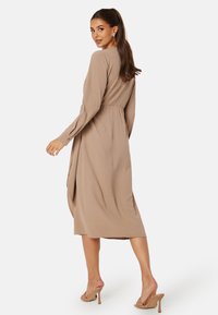 Beżowa sukienka z długim rękawem, z wcięciem w talii, o luźnej tkaninie i długości midi. Gładka tekstura i prosta sylwetka bez wzorów.