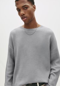 Junger Mann, der einen strukturierten hellgrauen Pullover, eine dünne silberne Halskette und beige Hose mit einem schwarzen Gürtel trägt, vor einem schlichten weißen Hintergrund.