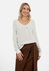 Weiße Bluse mit langen Ärmeln und V-Ausschnitt, vorne mit zwei goldenen Knöpfen versehen. Getragen mit braunen Hosen, die eine glatte Stofftextur zeigen.