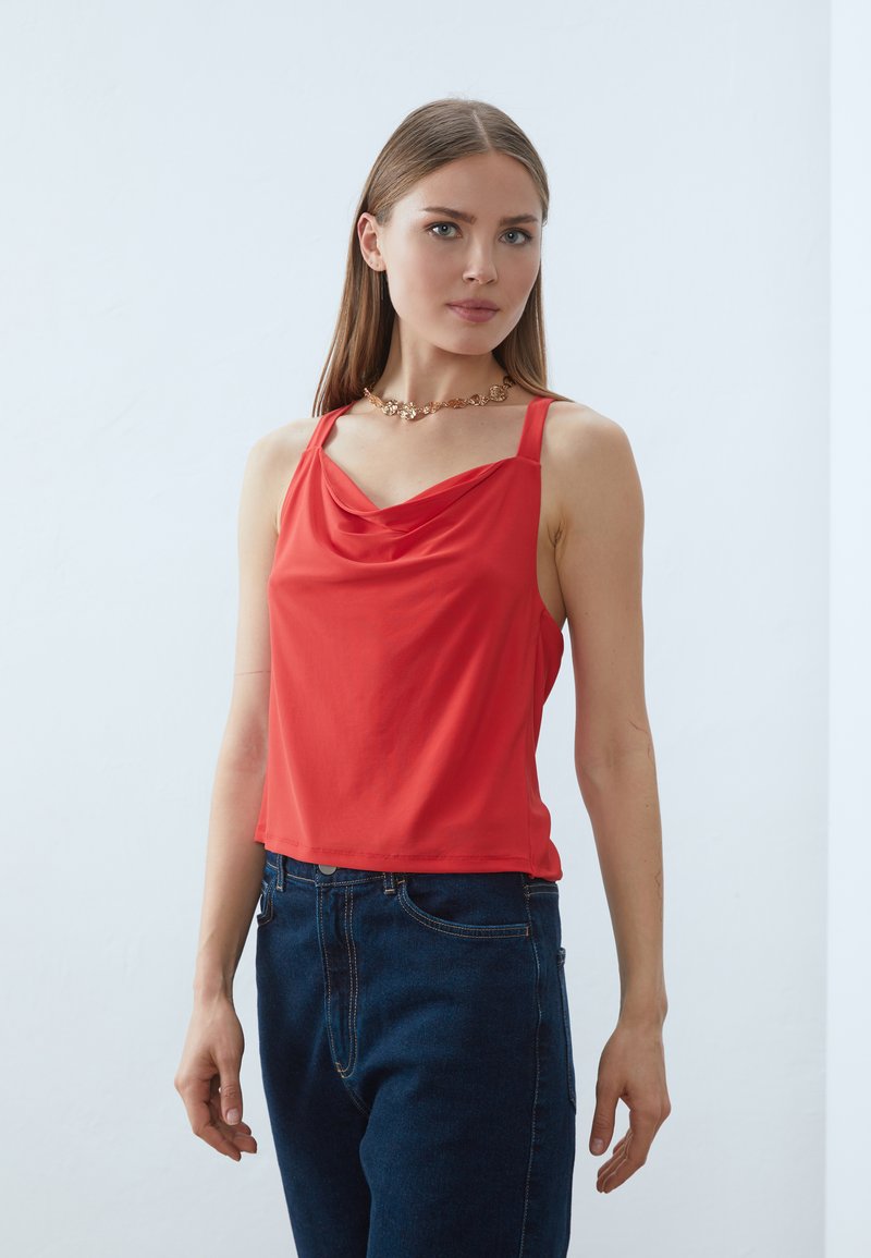 Top rosso senza maniche con scollo drappeggiato, realizzato in tessuto morbido. Abbinato a jeans in denim blu a vita alta.