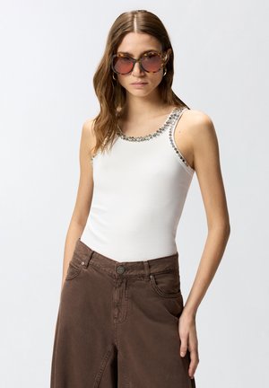 Γυναίκα που φοράει γυαλιά ηλίου με σχέδιο χελωνόστρακα, λευκό αμάνικο τοπ με διακόσμηση από στρας και καφέ ψηλόμεσο παντελόνι, με το χέρι στην τσέπη.