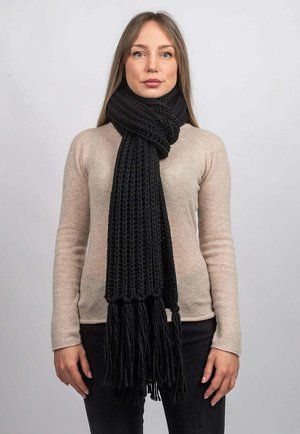 Sort strikket tørklæde med frynser, tykk tekstur og et chunky design, viklet om halsen. Båret over en lysefarvet sweater.