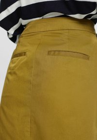 Vue arrière d'une jupe couleur moutarde avec deux poches fendues à l'arrière, portée avec un haut rayé noir et blanc.