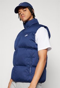 Gilet imbottito blu navy con colletto alto, chiusura con zip e tasche laterali. Presenta un logo Nike bianco sul petto. Indossato sopra una T-shirt bianca.