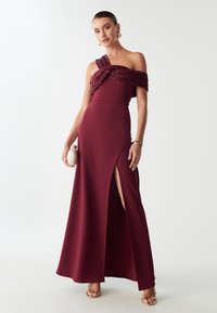 Bordo večernja haljina s asimetričnom naramenicom i nabranim detaljem od šljokica, koja se ističe visokim prorezom i oblinama koje ističu tijelo.