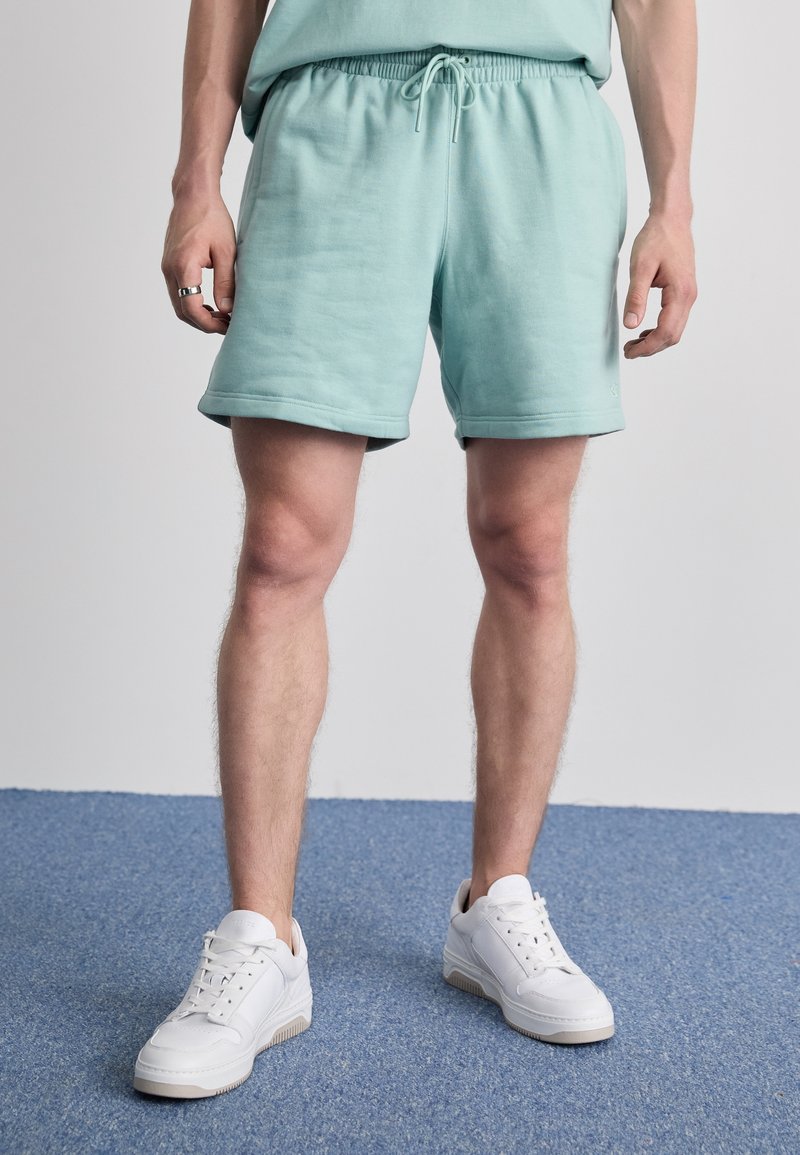 Minttu vihreät puuvillashortsit, joissa on joustava vyötärö ja naru. Yhdistettynä valkoisiin tennareihin, joissa on sileä pinta ja teksturoitu pohja.