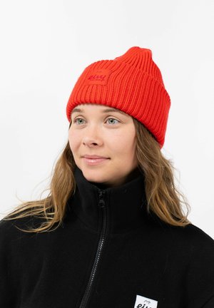 Rode gebreide beanie met een geribde structuur en een logo patch. Gedragen met een zwarte fleece jas met een hoge kraag en rits.