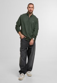 Dunkelgrüner Reißverschluss-Sweatshirt mit hohem Kragen, kombiniert mit legeren grauen Jeans und cremefarbenen Sneakers. Einfaches Design mit minimalen Details.