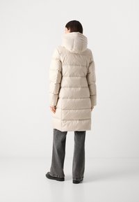 Beige dons jas met verticale quilting, capuchon en lengte tot halverwege de bovenbenen. Gekozen met grijze jeans en zwarte chunky schoenen. Soepele, gestructureerde stof.
