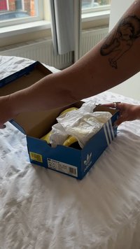 Caixa de sapatos azul da Adidas parcialmente aberta, revelando sapatos amarelos embrulhados em papel de seda branco, colocados sobre uma superfície branca texturizada.