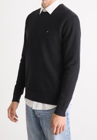 Schwarzer Strickpullover mit V-Ausschnitt, kombiniert mit einem weißen Hemd mit Kragen darunter. Verfügt über ein kleines Logo auf der Brust und lange Ärmel.