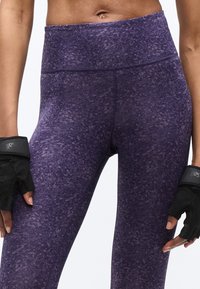 Gros plan sur une personne portant des leggings taille haute à motifs violets et des gants de fitness noirs, montrant le milieu du corps et le haut des jambes.