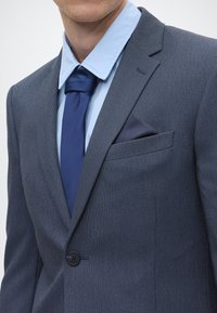 Casaco de fato azul-marinho com um tecido texturizado, fecho de um único botão e um bolso no peito contendo um lenço de bolso azul-escuro. Camisa e gravata azul-claro.