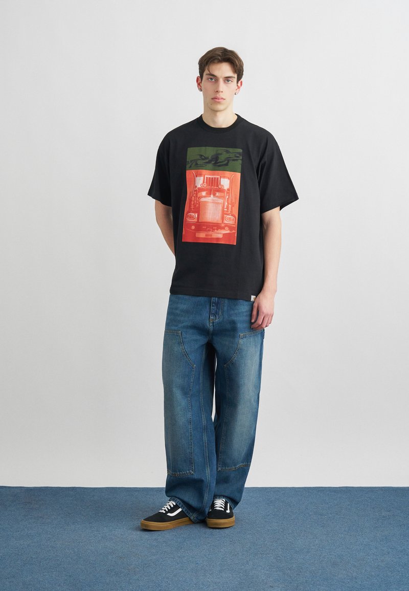 Joven con camiseta negra con un gráfico de un camión rojo y verde, jeans azules holgados y zapatillas negras, de pie sobre una alfombra azul.