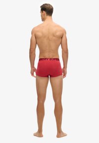 Boxer-briefs en coton rouge avec une ceinture élastique noire affichant le logo "SUPERDRY", présentés de dos sur un modèle pieds nus.