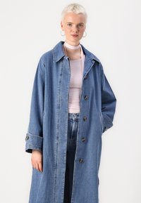Cappotto in denim lungo con vestibilità rilassata, colore azzurro chiaro, grande colletto, bottoni frontali e maniche arrotolate. Indossato sopra un top rosa.