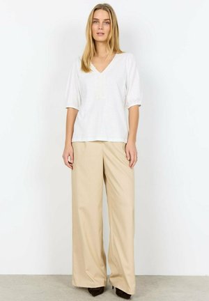 Vrouw met een witte V-hals blouse met borduursels, beige broek met wijde pijpen en donkere puntige schoenen, staand tegen een effen witte muur.