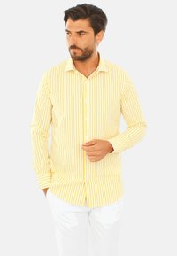 Camicia a maniche lunghe a righe gialle e bianche con colletto a punta. Materiale in cotone, design aderente e bottoni frontali aggiungono dettagli.