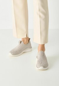 Világosszürke slip-on cipők texturált kötött felsőrésszel, fehér középtalppal és húzófülekkel a sarkán. Tejkrém színű nadrággal párosítva.