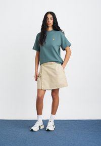 Πράσινο κοντομάνικο μπλουζάκι από βαμβάκι με κοντά μανίκια και κίτρινο λογότυπο, συνδυασμένο με μπεζ φούστα τύπου ντραπέ. Συμπληρώνεται με λευκές κάλτσες και αθλητικά παπούτσια.