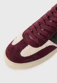 Baskets en daim bordeaux avec des accents en tissu texturé crème, une rayure en cuir noir et des lacets ronds. Présente une semelle en caoutchouc avec un motif triangulaire.