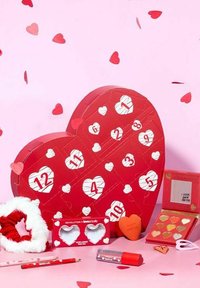 Röd hjärtformad presentlåda med numrerade flikar, omgiven av mindre hjärten dekorationer. Innehåller skönhetsprodukter, en spegel och en borste.