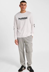 Wit shirt met lange mouwen met zwart "hummel"-logo, gecombineerd met grijze joggingbroek met logodetail. Model staat op een effen achtergrond.