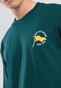 Tummanvihreä pitkähihaisempi t-paita, joka on valmistettu puuvillasta. Paidassa on keltainen ja valkoinen logo, jossa on lippumuotoilu ja teksti "New Port Sailors Yacht Club."