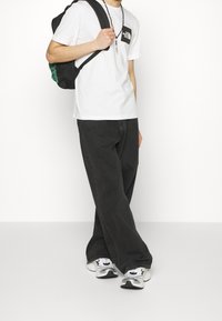 Person som bär en vit North Face T-shirt, svarta vida byxor, vita sneakers och bär en svart ryggsäck över ena axeln.