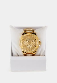 Zlaté Guess hodinky s kryštálovými akcentmi, chronografovými ciferníkmi a textúrovaným ciferníkom, vystavené na bielom vankúšiku v krabičke.