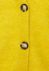Cárdigan de punto amarillo que presenta dos grandes botones redondos con un diseño moteado, una tela texturizada y una clásica parte delantera abotonada.