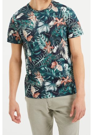 Homme portant un t-shirt à manches courtes à imprimé tropical foncé avec des feuilles vertes et des fleurs roses, associé à un pantalon beige clair.