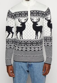 Stickad tröja med kvart zip, med en vit bakgrund prydd med svarta renar, snöflingor och geometriska mönster.