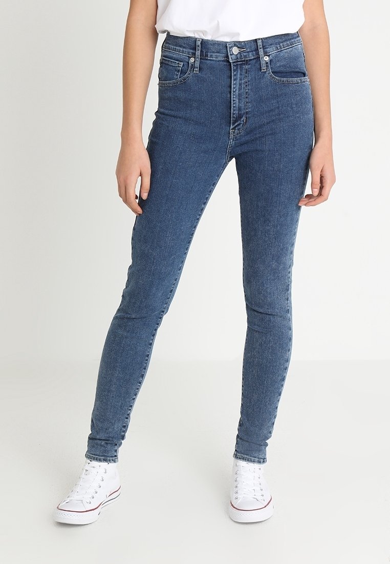 Jeans ajustados en denim azul oscuro, diseño de cintura alta, presentan un estilo tradicional de cinco bolsillos y herrajes de metal, combinados con zapatillas blancas.