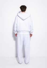 Jasnoszara bluza z dużym kapturem i ściągaczami, w zestawie z luźnymi spodniami dresowymi w tym samym kolorze, posiadającymi boczne kieszenie. Gładka, teksturowana tkanina.