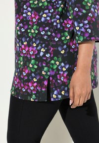 La tunica floreale presenta uno sfondo nero con fiori colorati in viola, rosa, verde e bianco. Orlo spaccato e maniche a tre quarti.