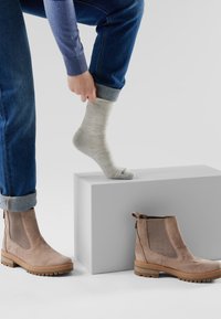 Stivaletti alla caviglia in suede color sabbia con pannelli laterali testurizzati, poggiati su una piattaforma grigia chiara. Una calza grigia chiara è tirata su su un piede. I pantaloni di denim sono arrotolati all'orlo.
