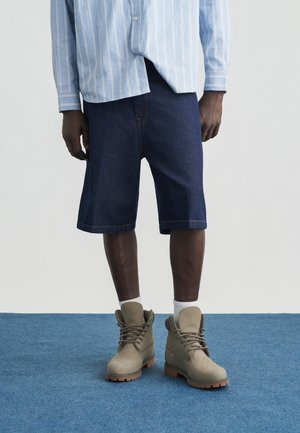 Isik, kes kannab siniseid teksapükse, helerohelist triibulist särki, valgeid sokke ja beežika värviga pitsitud saapaid, seismas sinisel vaibal.