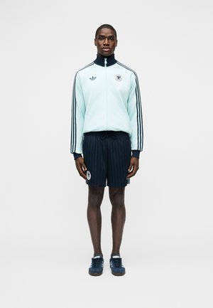 Egy férfi áll, előre néz, világoskék cipzáras dzsekit visel sötét csíkokkal, sötétkék rövidnadrágot és sportcipőt, egyszerű háttér előtt.