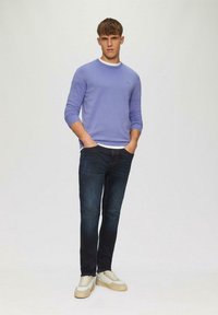 Leichter lila Pullover mit Rundhalsausschnitt, über einem weißen Hemd getragen. Dunkelblaue Jeans und beige Sneakers runden das lässige Outfit ab.