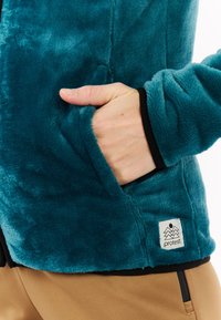 Melsvai mėlynas polaras su minkšta tekstūra, priekinė kišenė ir juodas apvadas. Apatinėje dalyje yra stačiakampis logotipo užrašas.