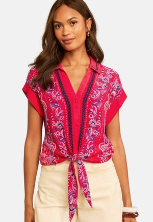 Frau trägt eine kurzärmelige Bluse mit rotem Blumenmuster, die in der Taille gebunden ist, und hellbeige Hose.