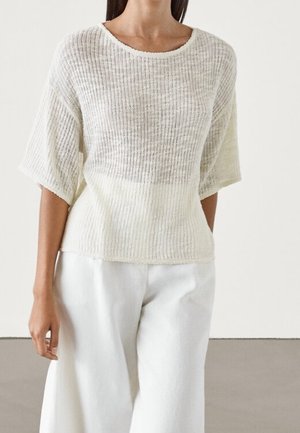 Vrouw draagt een losse, semi-transparante, geribde crèmekleurige trui met ellebooglange mouwen en wijde witte broek, staand tegen een effen achtergrond.