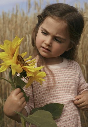 Kind hält eine große Sonnenblume und trägt ein rosa kariertes gemustertes Kleid. Im Hintergrund sind hohe Weizenfelder zu sehen. Die Sonnenblume hat leuchtend gelbe Blütenblätter und Samen.