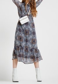 Vrouw in een lange bloemenjurk, met witte enkellaarsjes en een witte schoudertas, staande met opgetilde armen tegen een effen achtergrond.