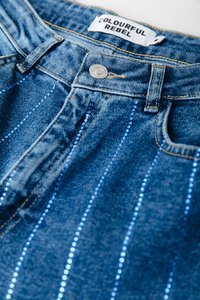 Jeans de mezclilla con una cintura de altura media, que presentan rayas verticales azules hechas de cuentas y un cierre de botón metálico. La etiqueta dice "COLOURFUL REBEL."