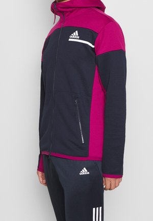 Homme portant une veste zippée Adidas noire et magenta avec un pantalon Adidas noir assorti avec des bandes blanches et des logos.
